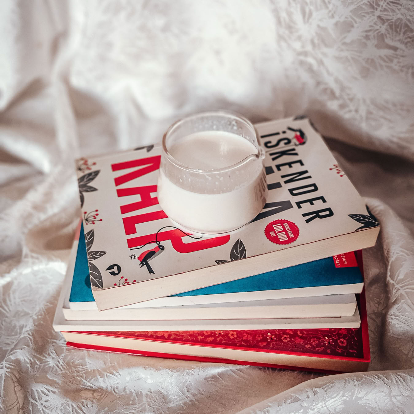 A stack of colorful books with a glass of milk on a draped fabric background.