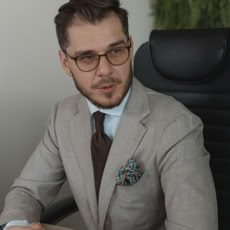 A professional man in a suit and glasses sits confidently at a desk in a modern office.